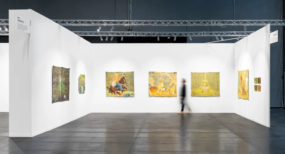 A Perrotin booth presents a series of luminous, textured paintings in greens, yellows and oranges as a blurred visitor walks through the space.