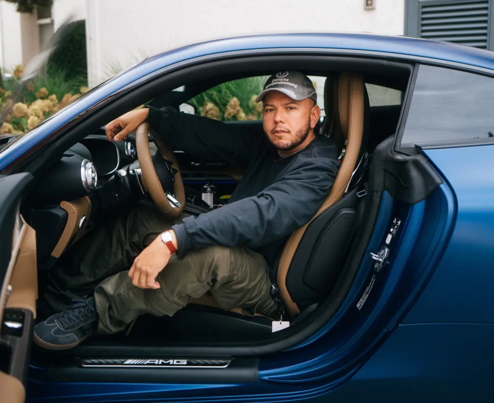 A man wearing a black sweatshirt and hat sits in the driver's seat of a blue Mercedes-AMG with the door open and looks into the camera.