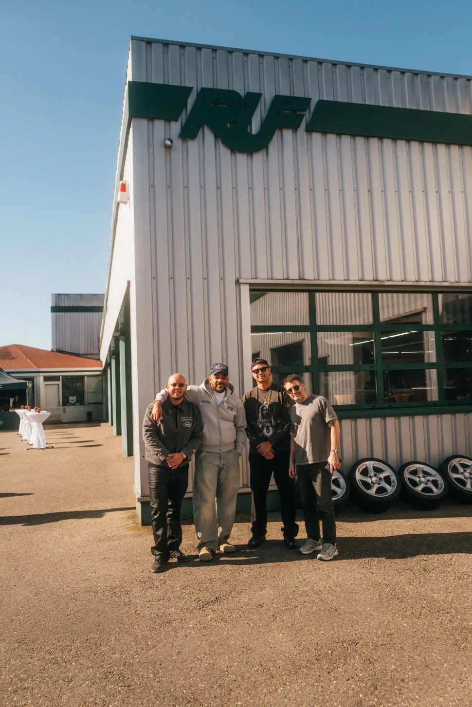 Four men posed for a photo outside an industrial building where a large green RUF logo was hung and several wheels lined up next to it.