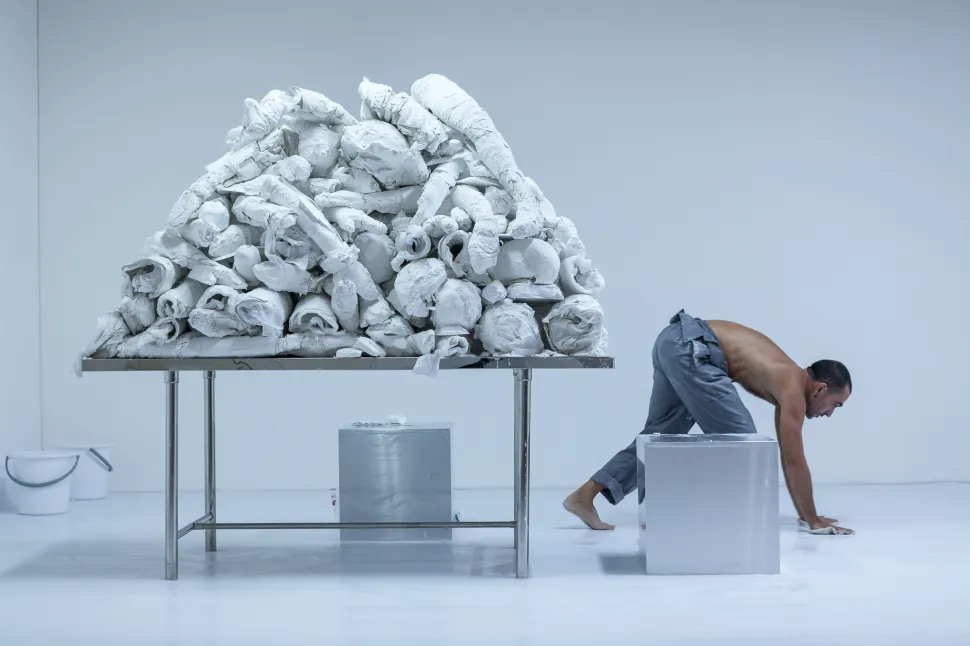 A performer moves beside a white mound of sculptural forms in Yannis Pappas’s Telephus, staged in a stark white space.