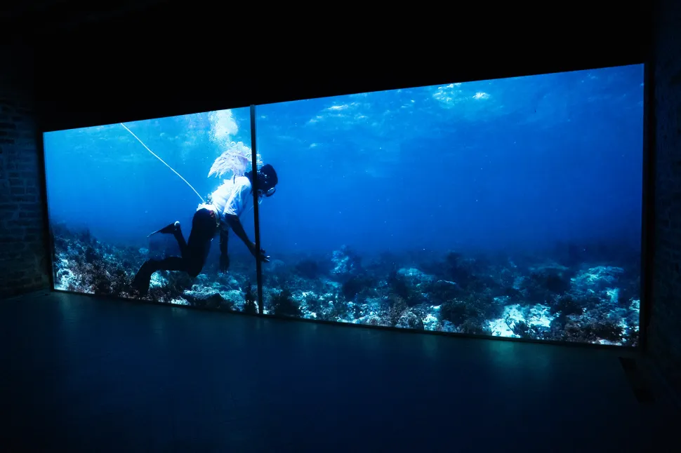 A large video projection by Martha Atienza shows a figure moving underwater across a blue ocean scene in a darkened exhibition space.