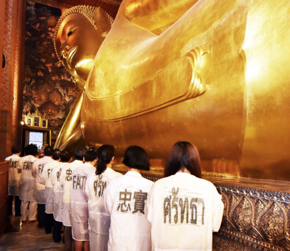 A group of people in white garments stand before a monumental reclining golden Buddha in Jitsing Somboon’s Faith Way.