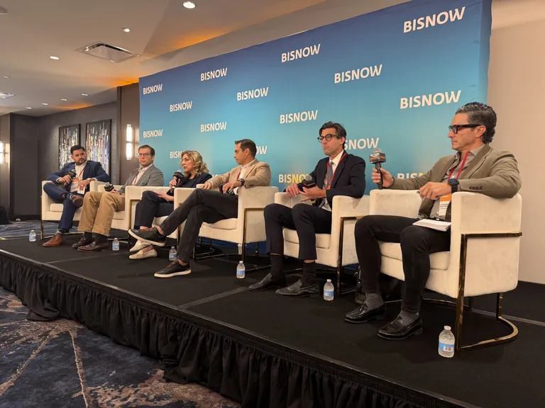 A panel of six people sat on stage at a conference, each holding a microphone, with a "BISNOW" background and bottled water at his feet.