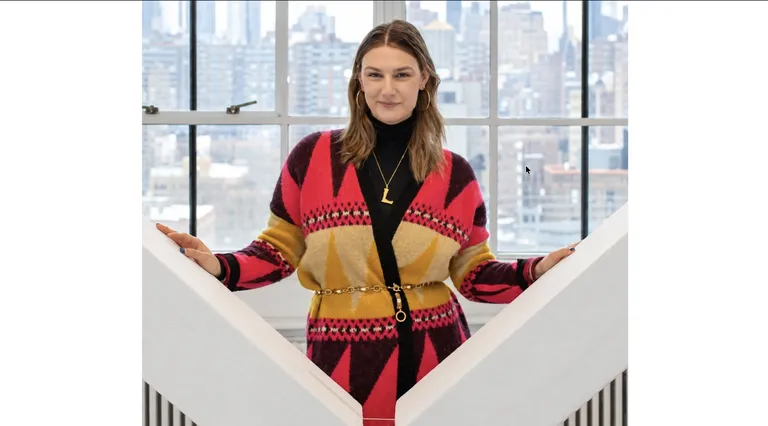 A man is smiling in front of a window overlooking a cityscape, wearing a geometric patterned cardigan with a gold chain belt.