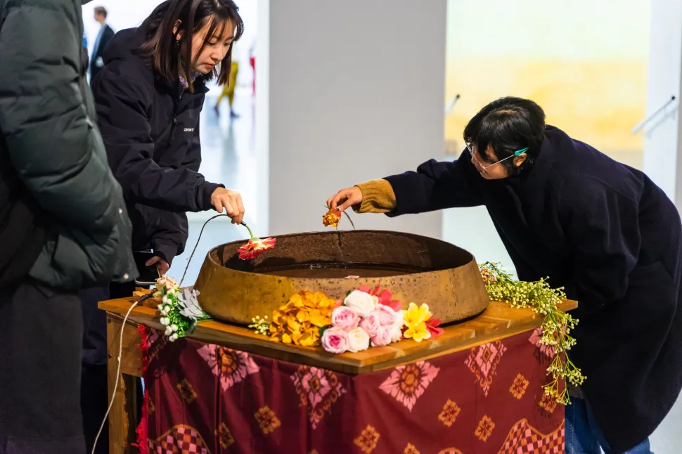An interactive sound installation by Samboleap Tol shows visitors placing flowers into a water-filled gong surrounded by artificial and fresh flowers.