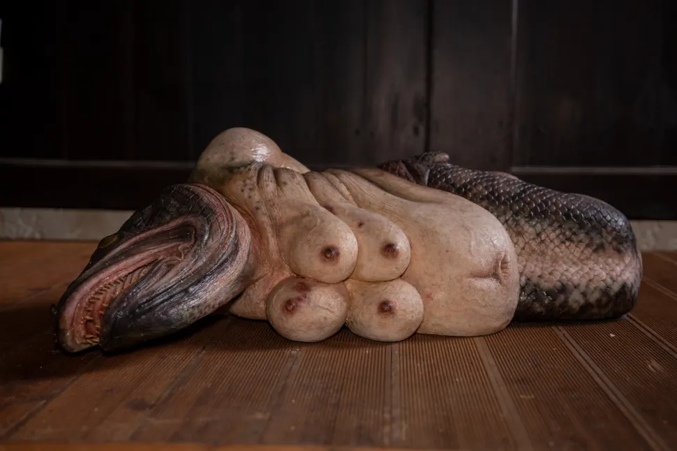 A mixed-media sculpture by Parada Wiratsawee combines a fish-like body with human anatomical forms on a wooden floor.