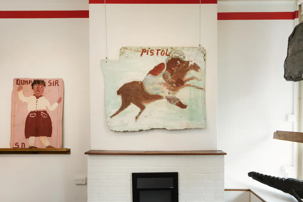 A gallery installation of Sam Doyle’s paintings includes a horse and rider beneath the word “Pistol” and a portrait of a standing figure titled Dump Sir, both painted on weathered, irregular surfaces.