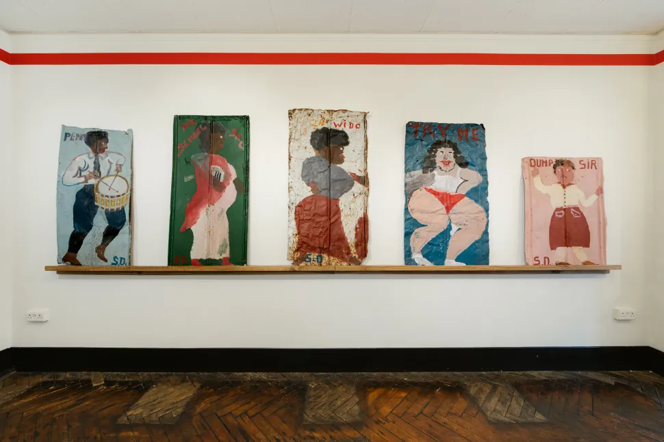 A row of five colorful paintings by Sam Doyle depicting expressive human figures, including a drummer and several women, rests on a wooden shelf against a white gallery wall.