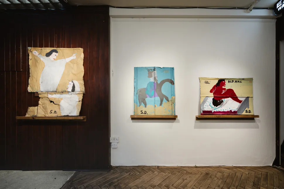 An installation view of three paintings by Sam Doyle depicting expressive human figures, a horseback rider and a red figure labeled “Old Hag,” displayed on wooden shelves against contrasting dark and white gallery walls.