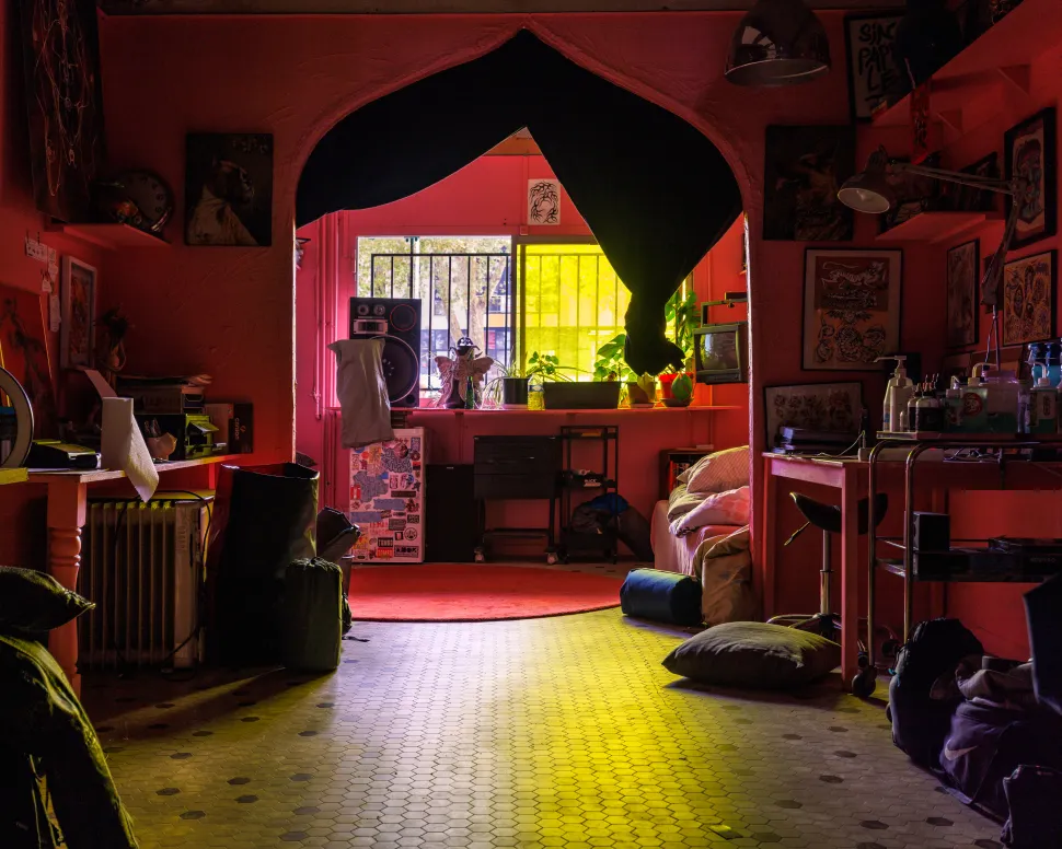 A pink-walled artist’s living and working space at Le Wonder, filled with framed artworks, personal objects and furniture, with a large black curtain hanging over an arched opening and sunlight filtering through colorful windows.