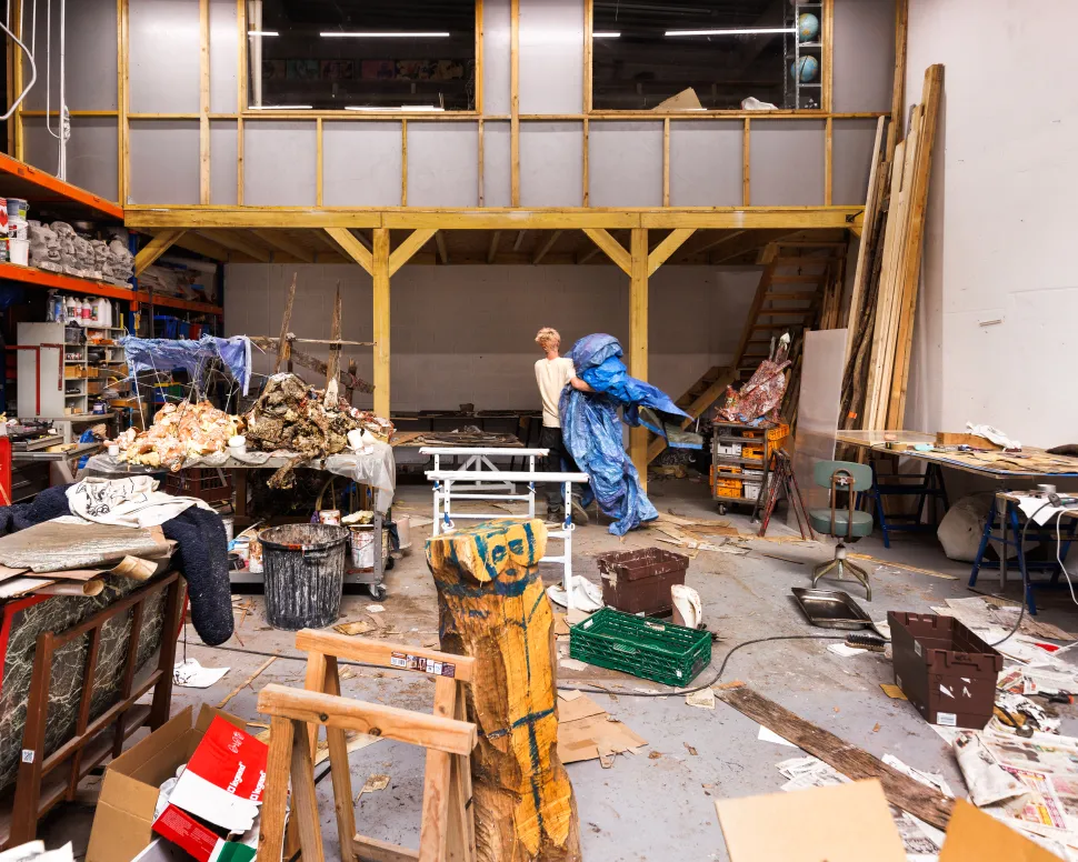 An artist carrying a large blue tarp through a spacious industrial workshop at Le Wonder, surrounded by sculptures, construction materials, tools and worktables beneath a wooden mezzanine.