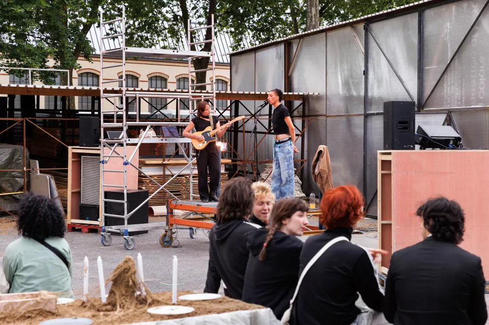A guitarist and singer performing on a makeshift outdoor stage at Le Wonder, with audience members gathered around tables in the foreground and industrial structures framing the courtyard.