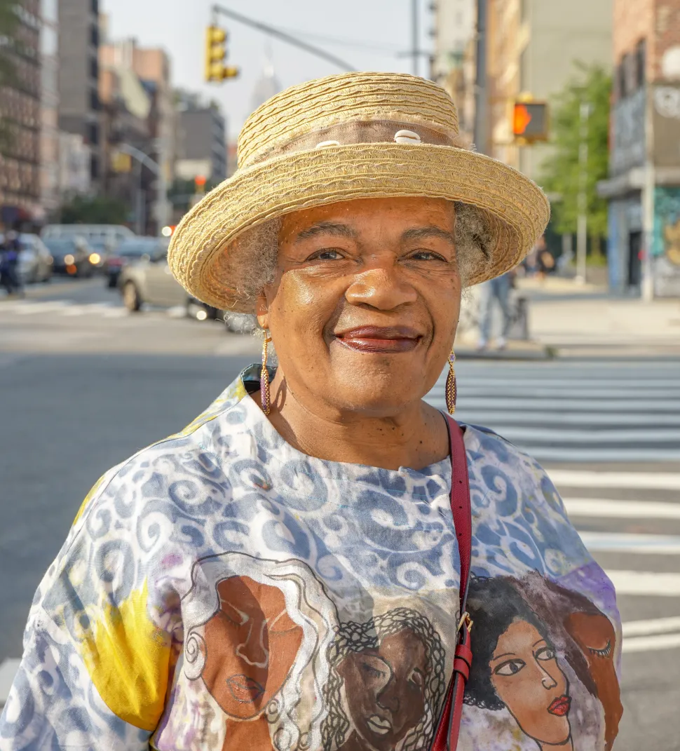 A portrait of Dindga McCannon smiling on a New York street, wearing a wide-brimmed straw hat and a colorful garment printed with women’s faces.