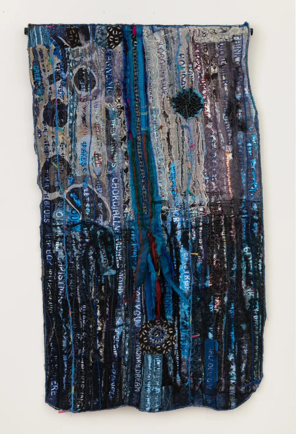 A mixed-media quilt by Dindga McCannon composed of layered blue, black and gray fabric strips, stitched text and circular decorative elements.