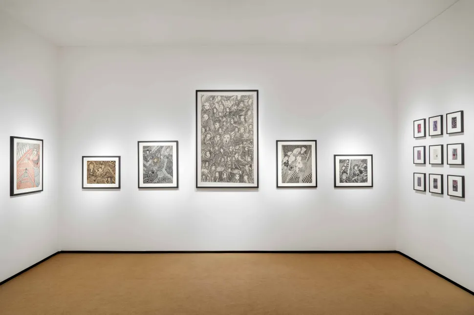 An exhibition installation at Frieze features a selection of framed, densely detailed figurative drawings arranged across white gallery walls, including a large central composition populated by numerous overlapping faces and figures.