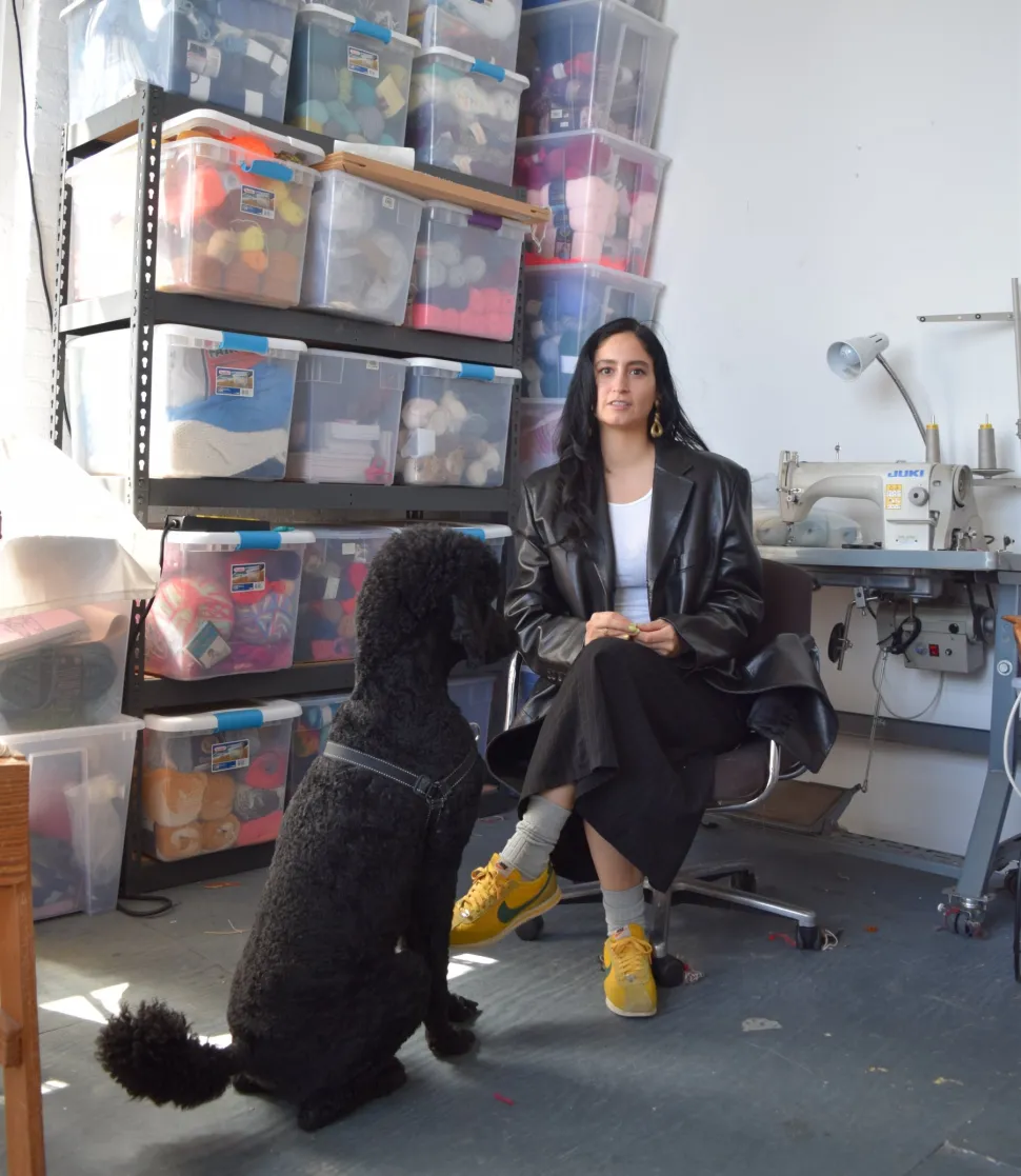 An artist sits in her studio with a large black dog next to her, surrounded by shelves of colorful yarn and textile materials, and an industrial sewing machine to the right.