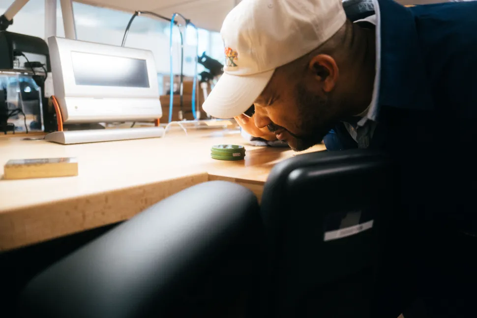 In the workshop, a man in a white hat leaned on the workbench to inspect a small part next to precision equipment.