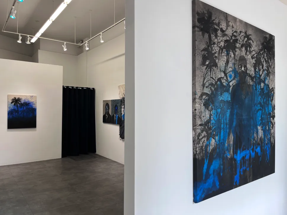A gallery installation view showcases several paintings in dark, blue and black colors, displaying images of palm trees and shadowed figures against white walls.