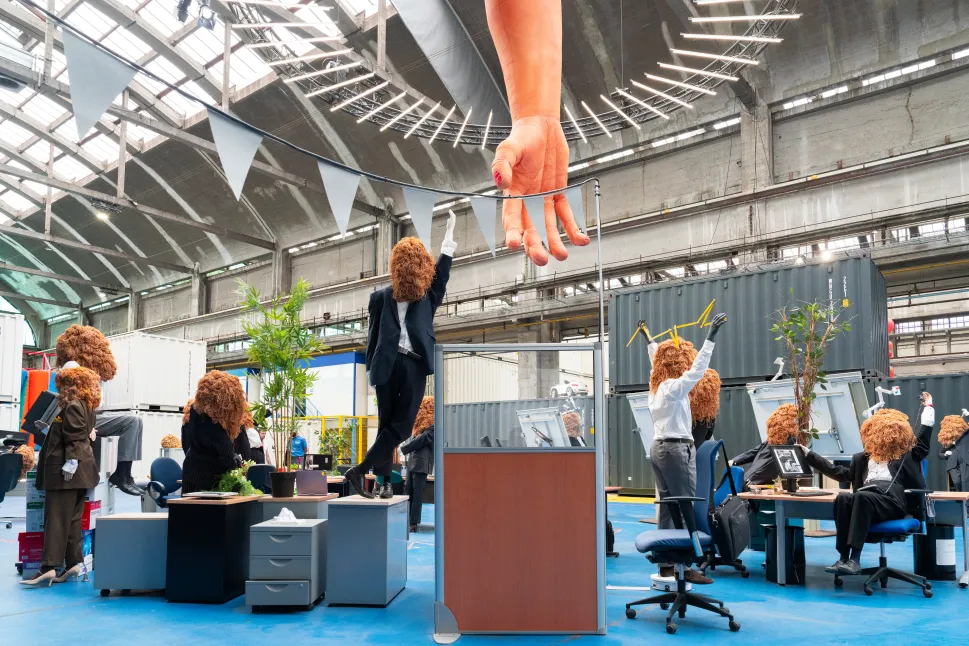 This is an art installation in an office with a giant orange hand with a spaghetti head humanoid on it