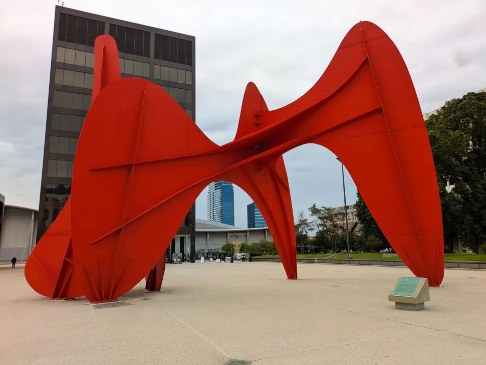 A monumental red abstract steel sculpture rises in a plaza surrounded by modern buildings in Grand Rapids.