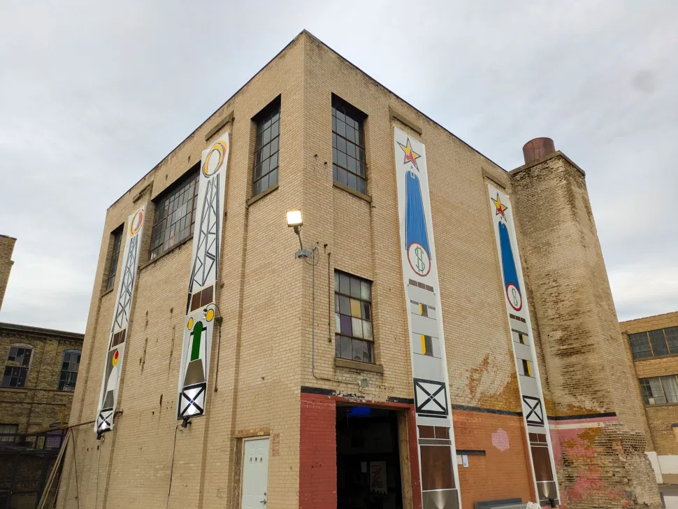 An industrial brick building in Grand Rapids is decorated with tall vertical banners featuring colorful geometric and symbolic designs.