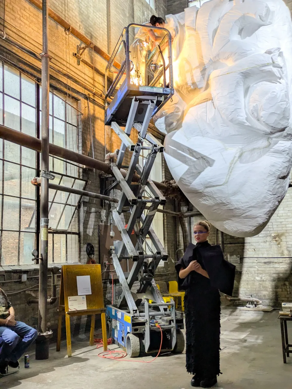 A woman in black stands beside a scissor lift while another person works above her on a monumental suspended white sculpture in an industrial space.