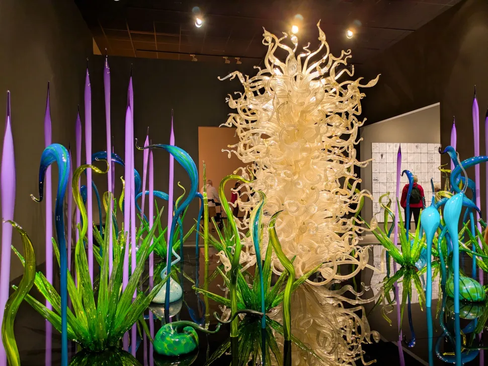 A colorful glass installation combines tall purple spikes, curling green and blue forms and a dense central mass of clear glass tendrils inside a dark gallery.