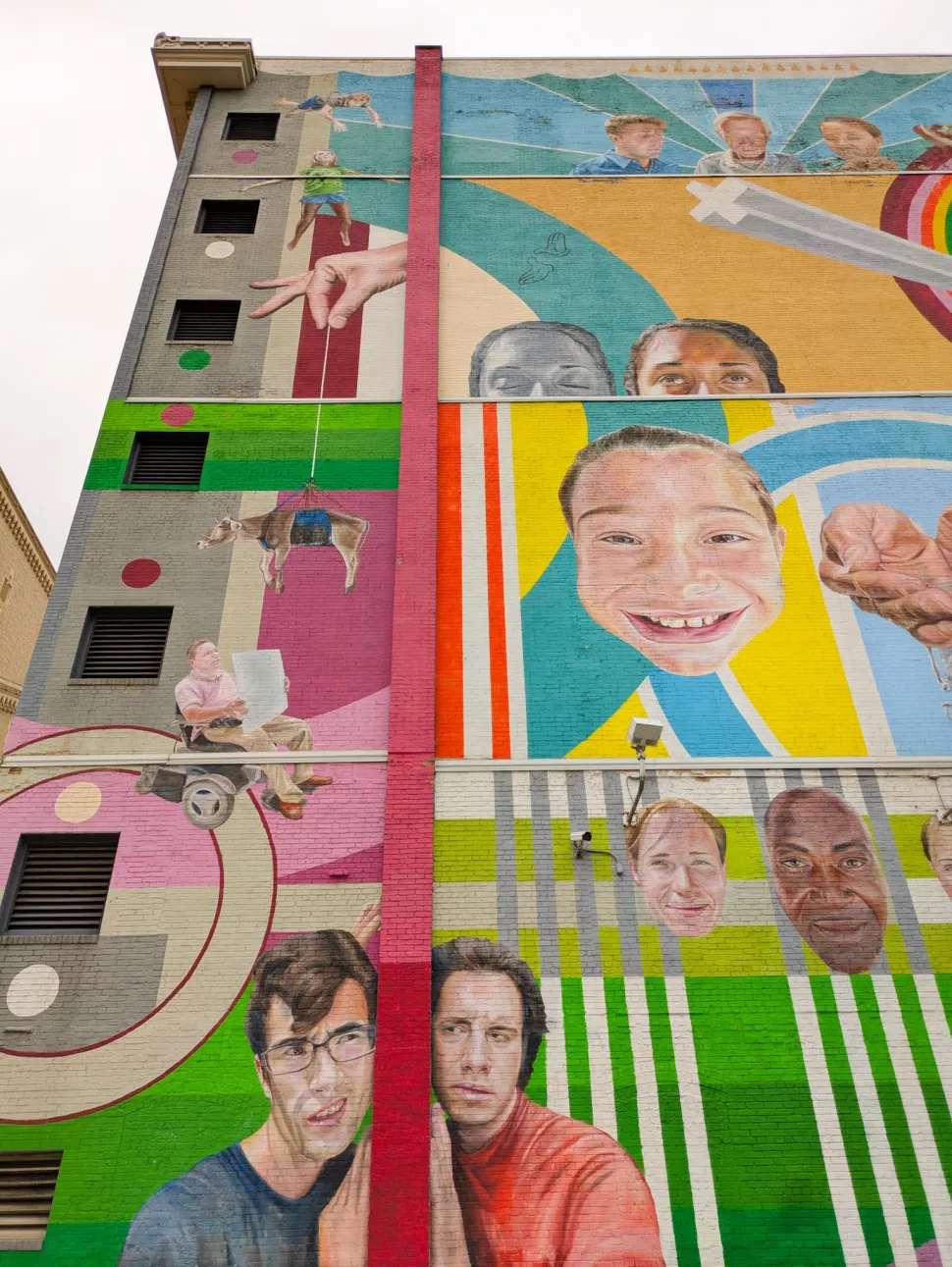 A large multistory mural covers the side of a building with brightly colored geometric patterns, painted portraits, hands and suspended figures.