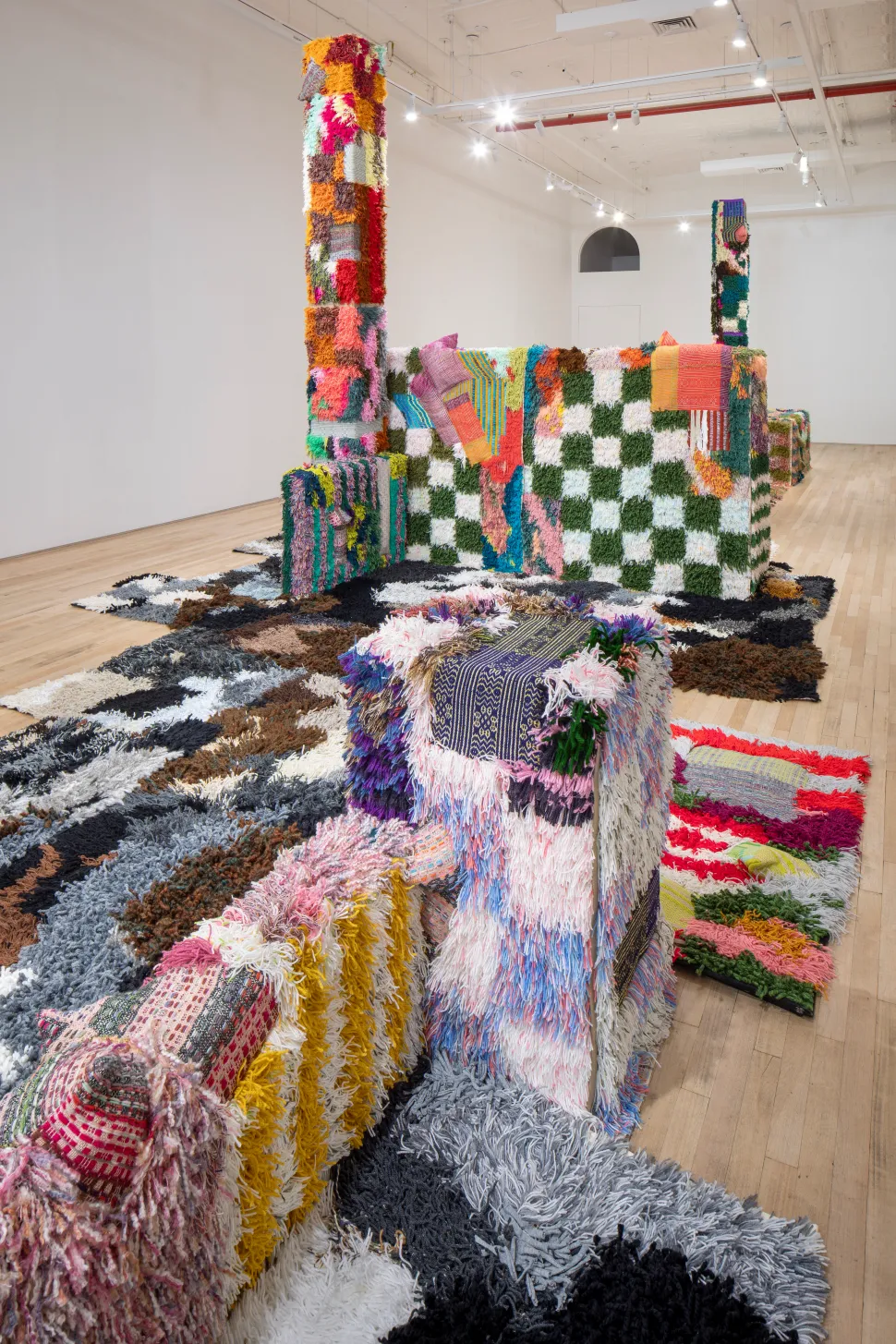 Installation views of Sarah Zapata’s brightly colored handwoven and textile-covered architectural forms are spread throughout the gallery floor, with tall columns and layered furry surfaces creating an immersive environment.
