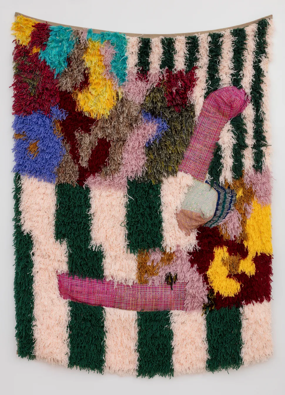 The wall-mounted textile consists of furry green, cream, red, yellow and blue sections intertwined with patterned woven forms that protrude from the surface.