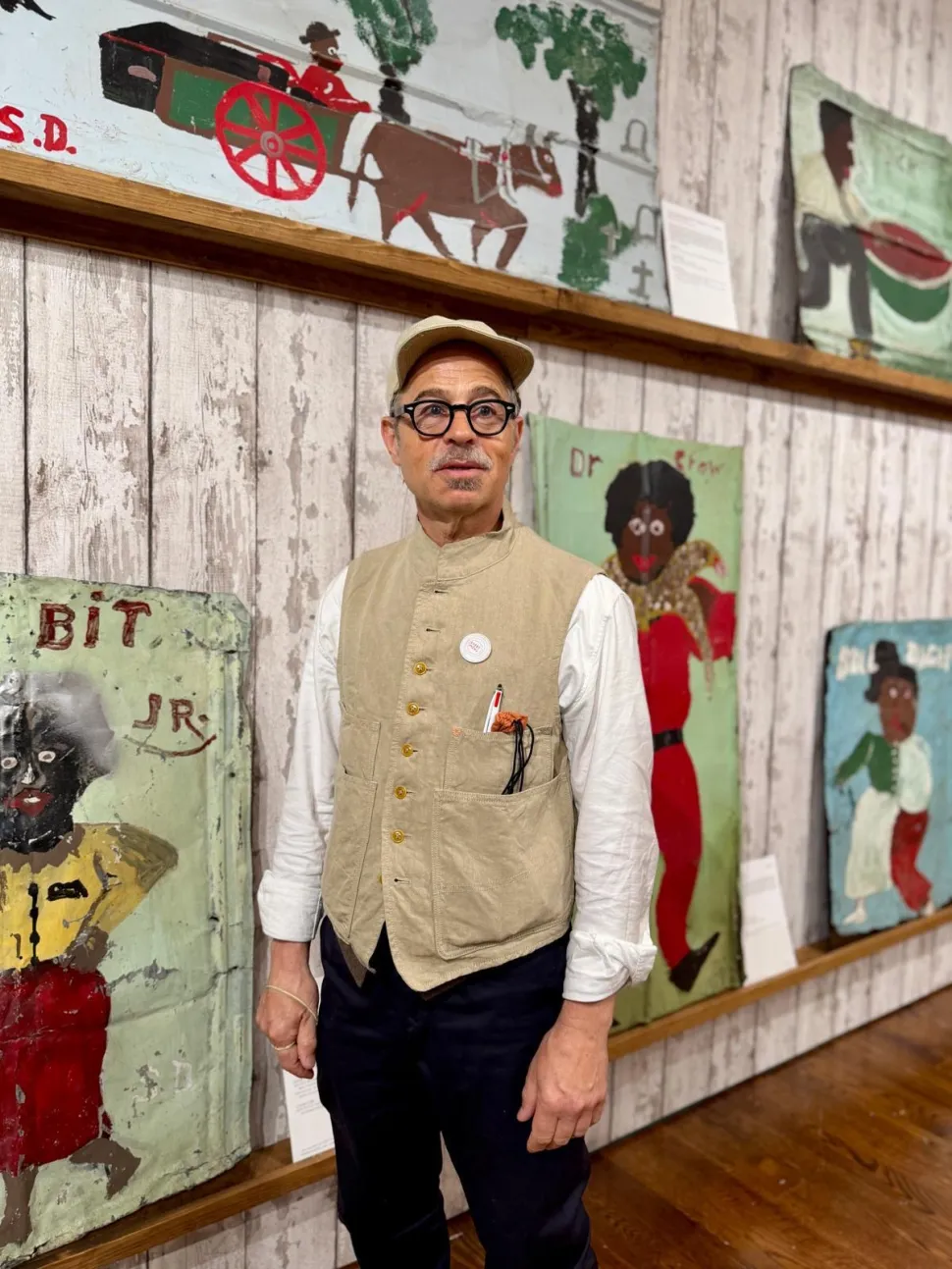 A man wearing glasses, a beige cap and a tan vest stands in front of a display of Sam Doyle’s colorful paintings of figures and scenes from African American life at The Gallery of Everything.